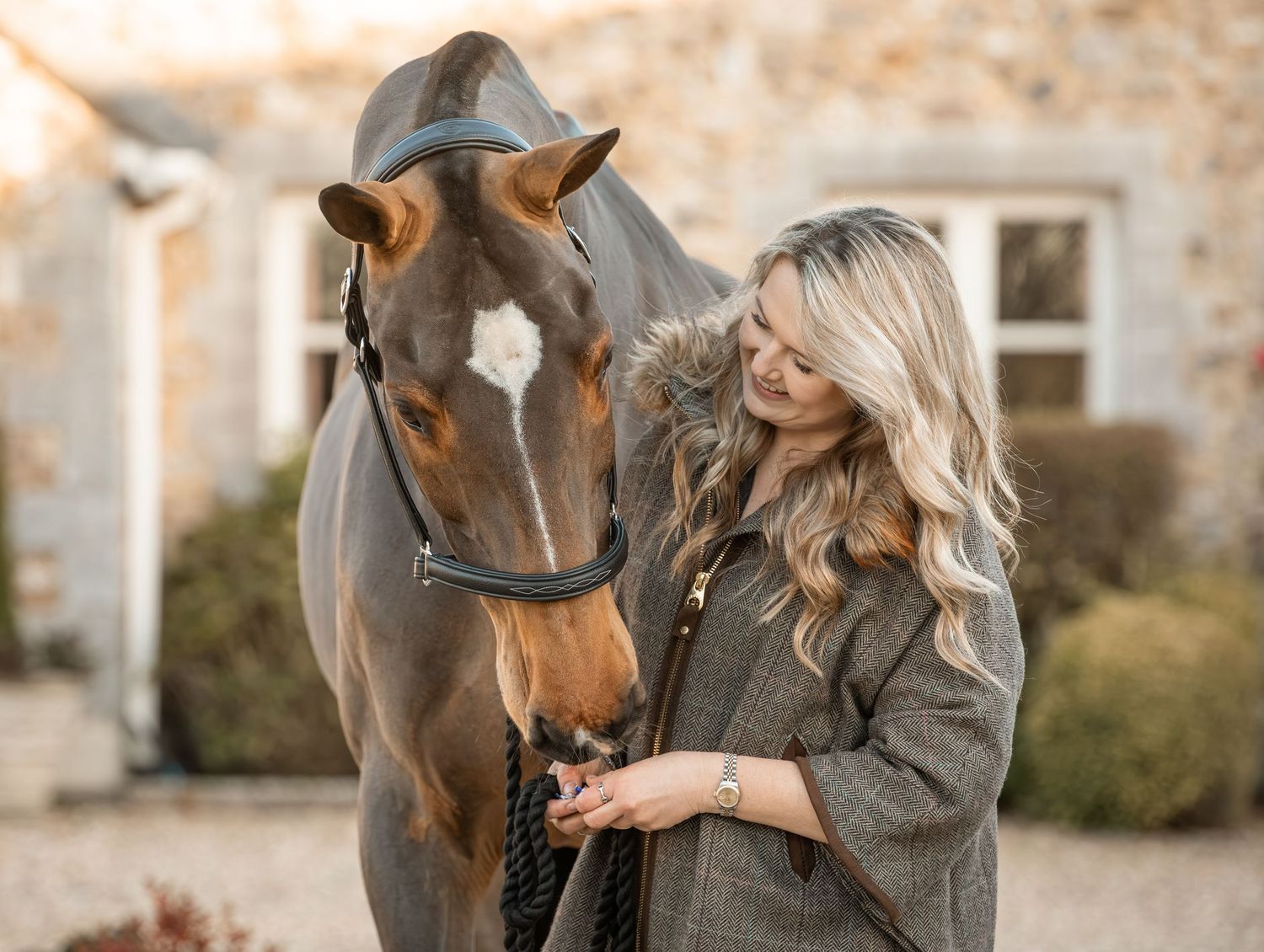 A brown horse and rider share a tender moment in front of a rustic stone wall during an equestrian photoshoot.