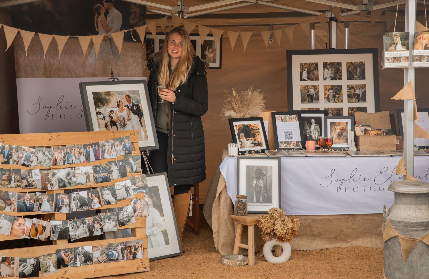 Woman standing at an outdoor market stall displaying framed family photos and prints for sale.