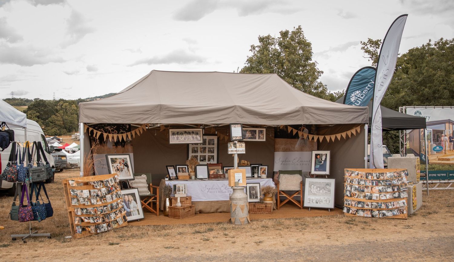 Outdoor art vendor booth with tent displaying framed artwork and prints at an open-air market or festival.