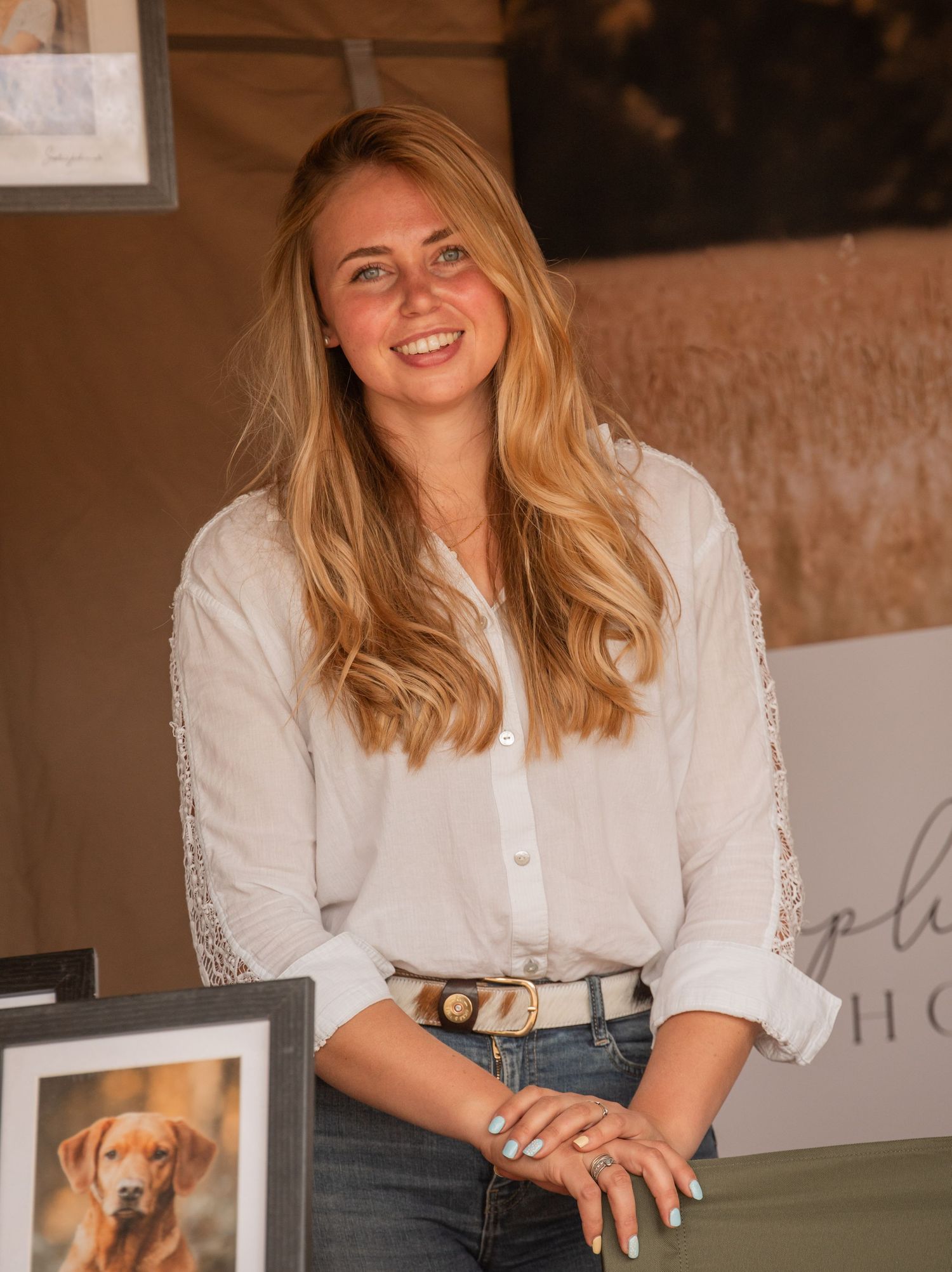 Smiling blonde woman in white blouse and jeans stands beside a framed dog portrait at an art display booth.