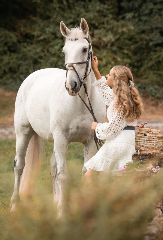 A dreamy outdoor photoshoot features a white horse and someone in a flowing white lace dress in a natural setting.