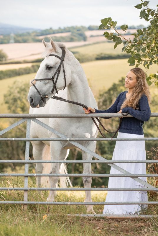 A majestic white horse poses against a scenic countryside backdrop with rolling hills and wooden fencing.
