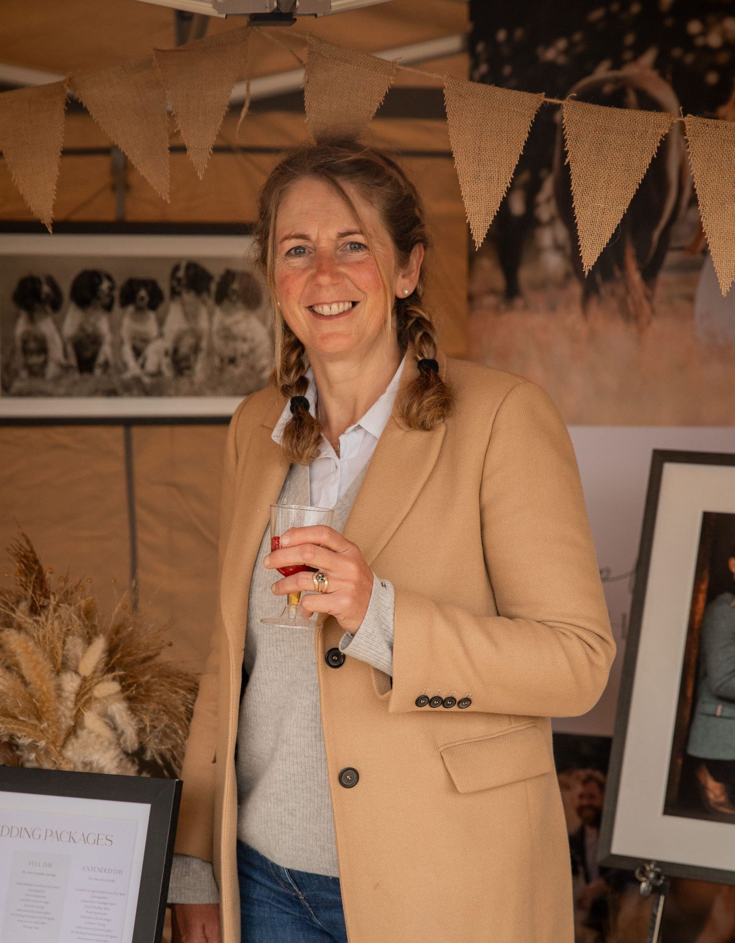 Woman in camel blazer smiling at an indoor event, holding a drink with bunting decorations in the background.