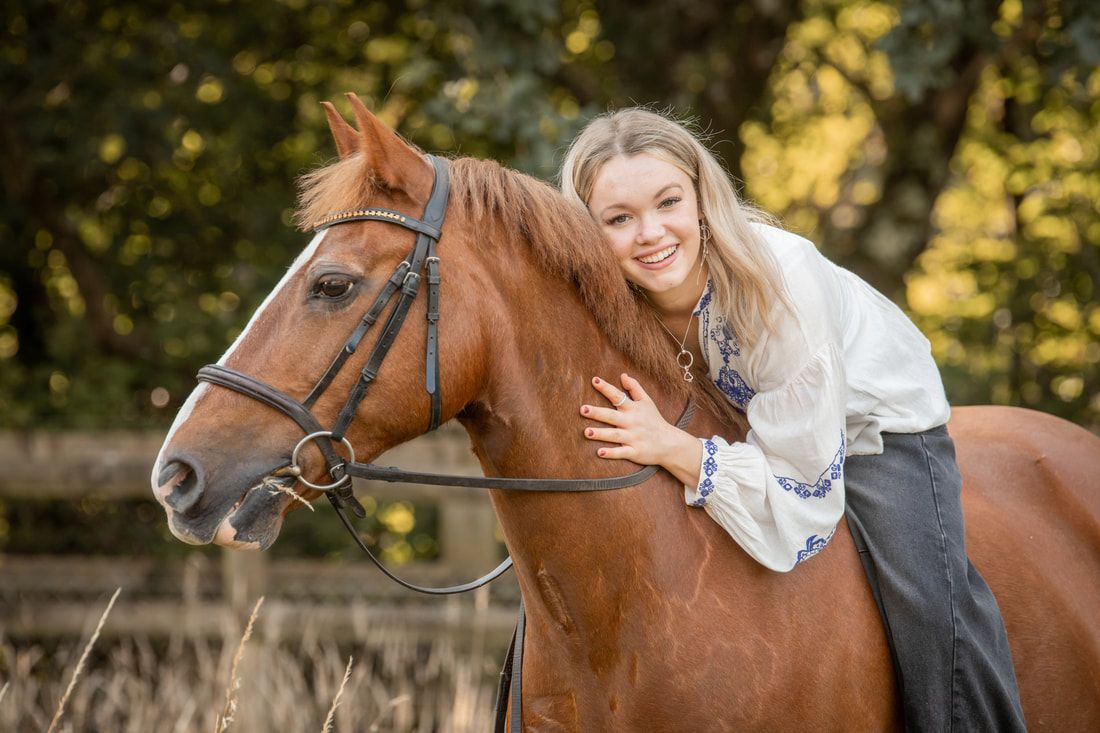 A chestnut horse and rider pose together in a sunlit outdoor setting with trees in the background.