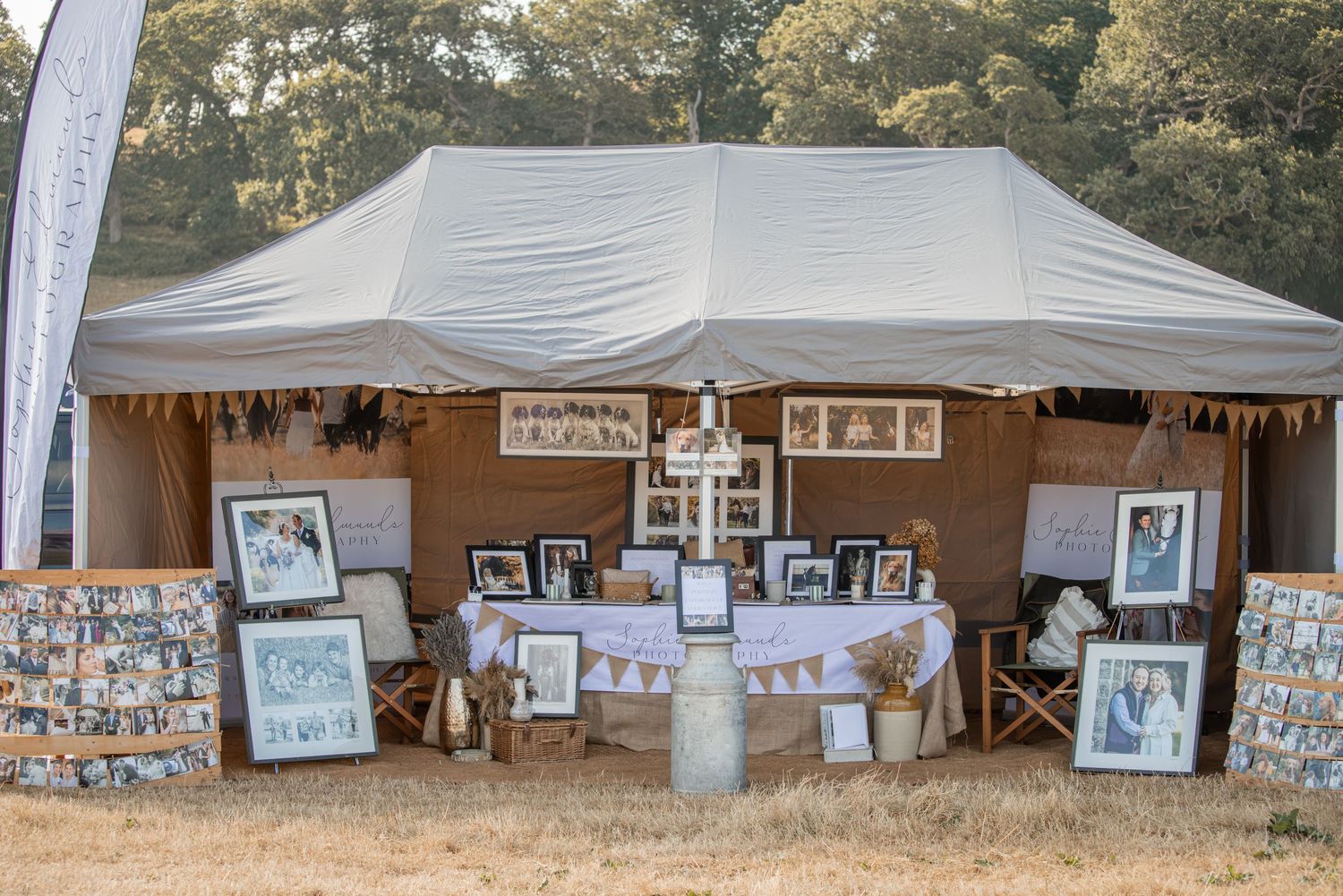 Rustic outdoor wedding display booth with framed photos, chandelier, bunting, and burlap decor under a large tent.