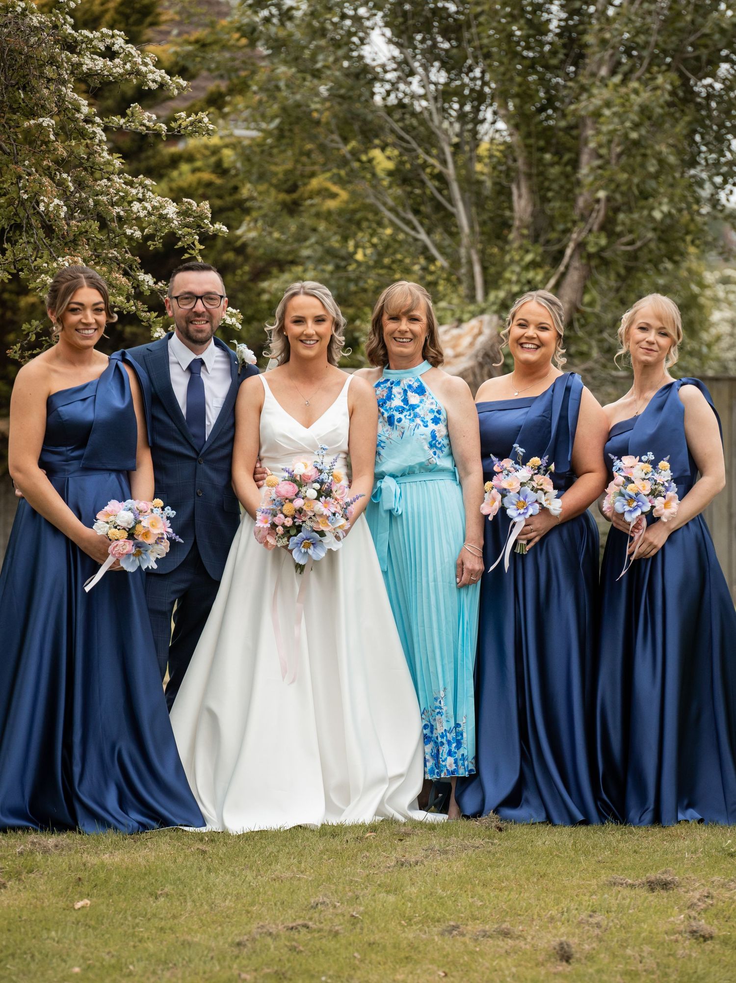 Bride in white gown poses with bridesmaids in navy blue dresses and groomsman in navy suit outdoors.
