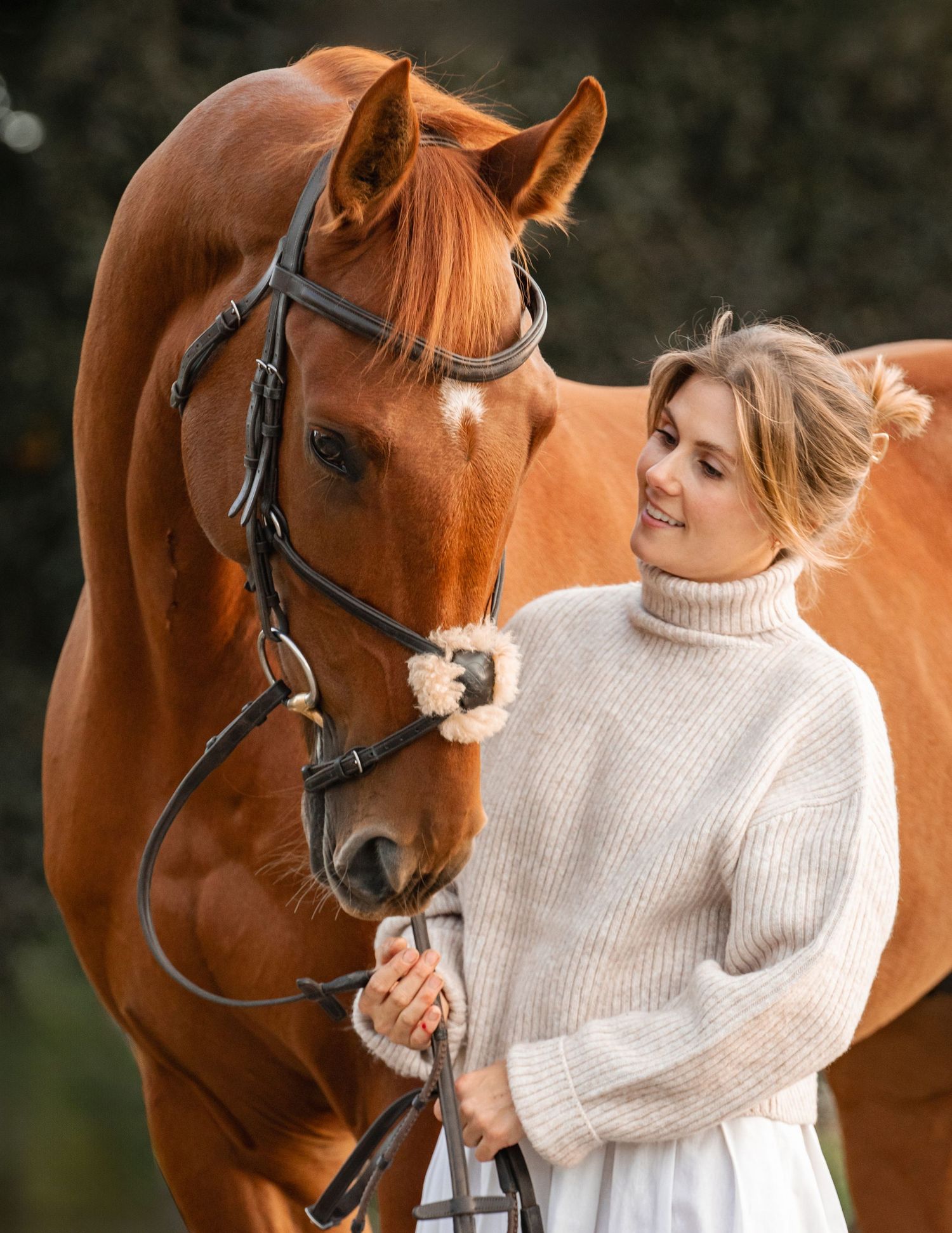 A woman in a cream sweater shares a tender moment with a chestnut horse wearing a bridle against a dark background.