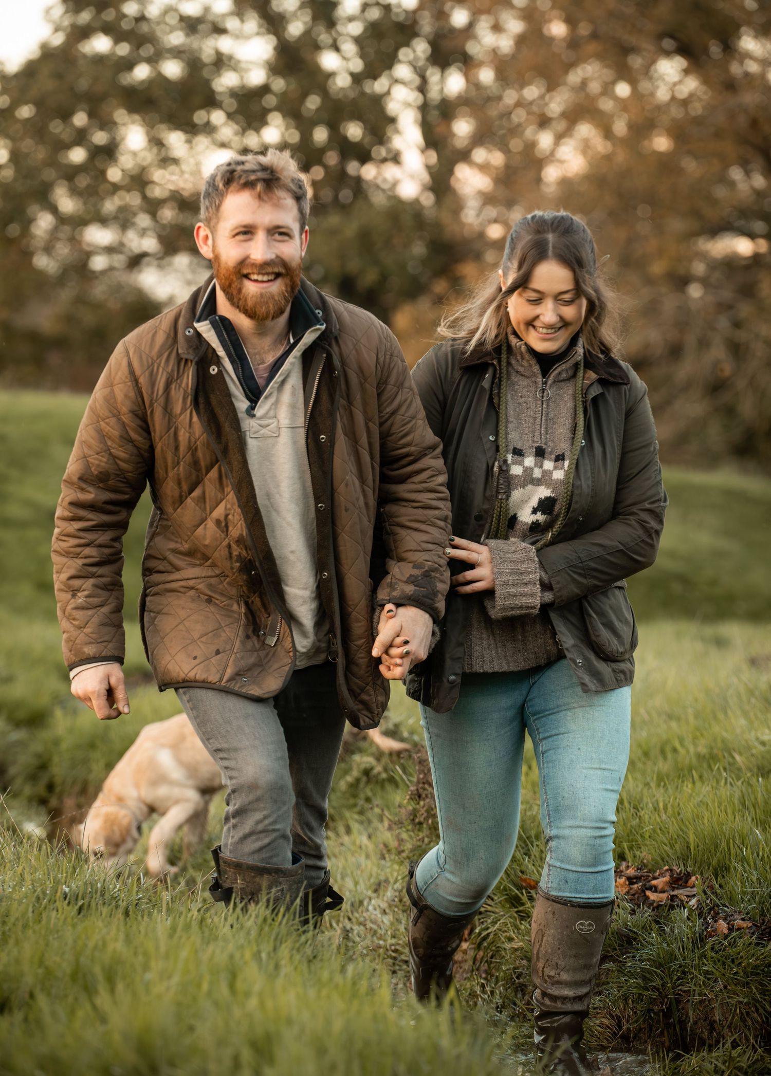 A couple in casual attire sits together on grassy hillside during golden hour with their dog in autumn setting.