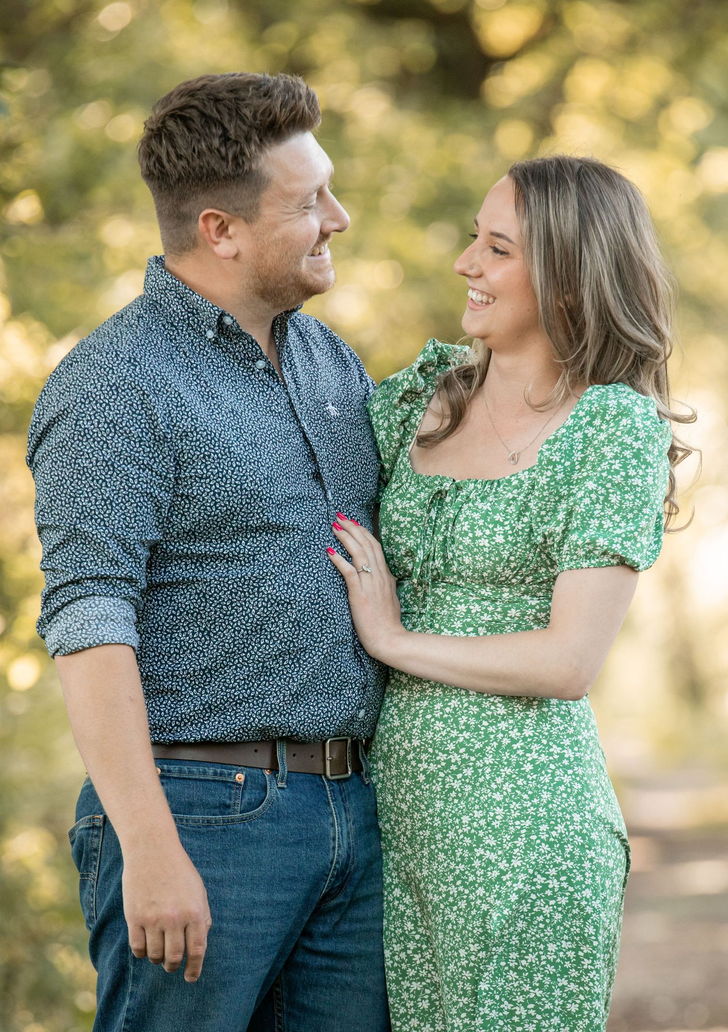 A couple shares a joyful moment outdoors, with her wearing a green floral dress and him in a blue patterned shirt.