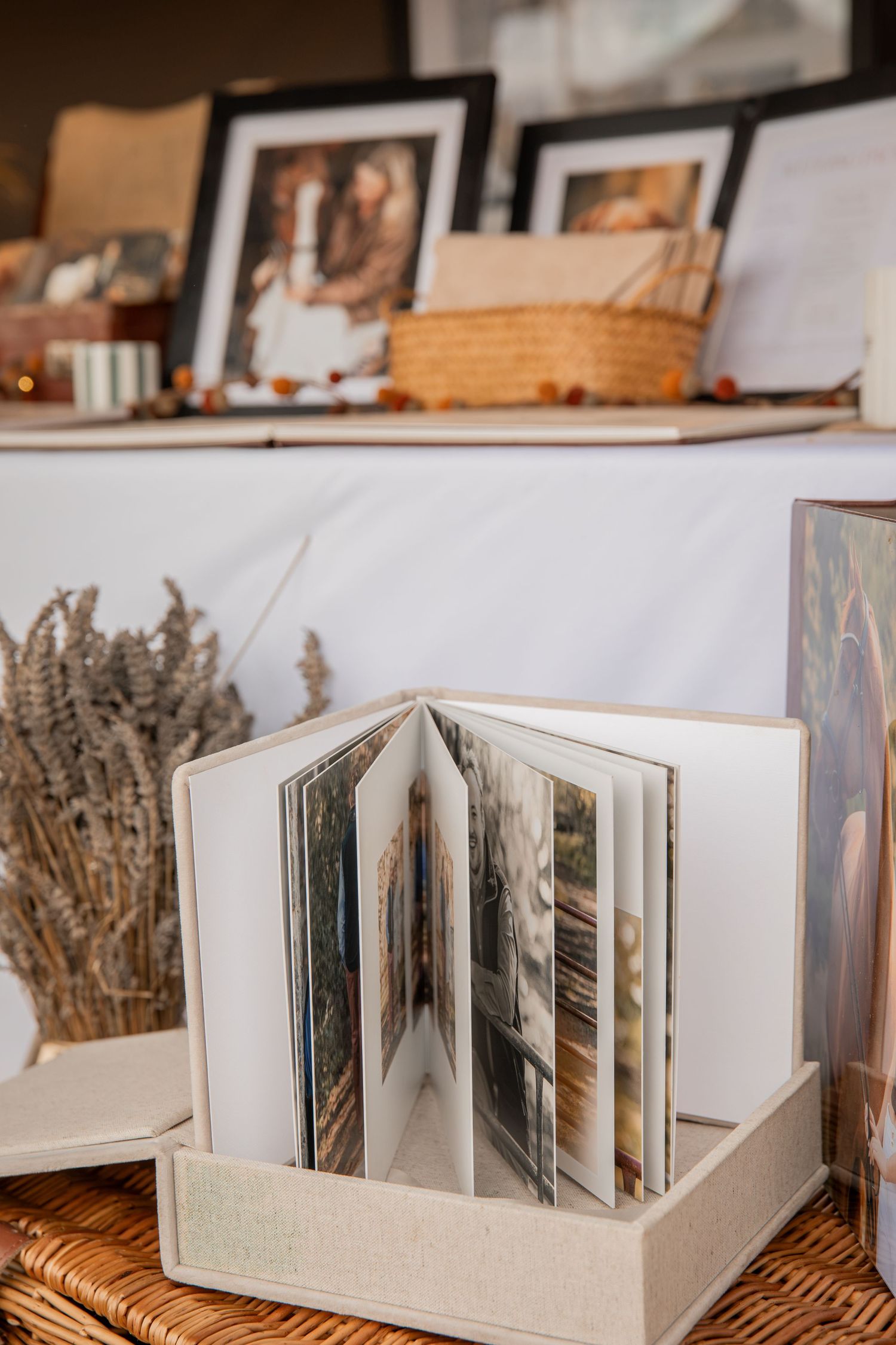 Open photo book displayed in a linen box with framed pictures and wicker basket on shelf in background.