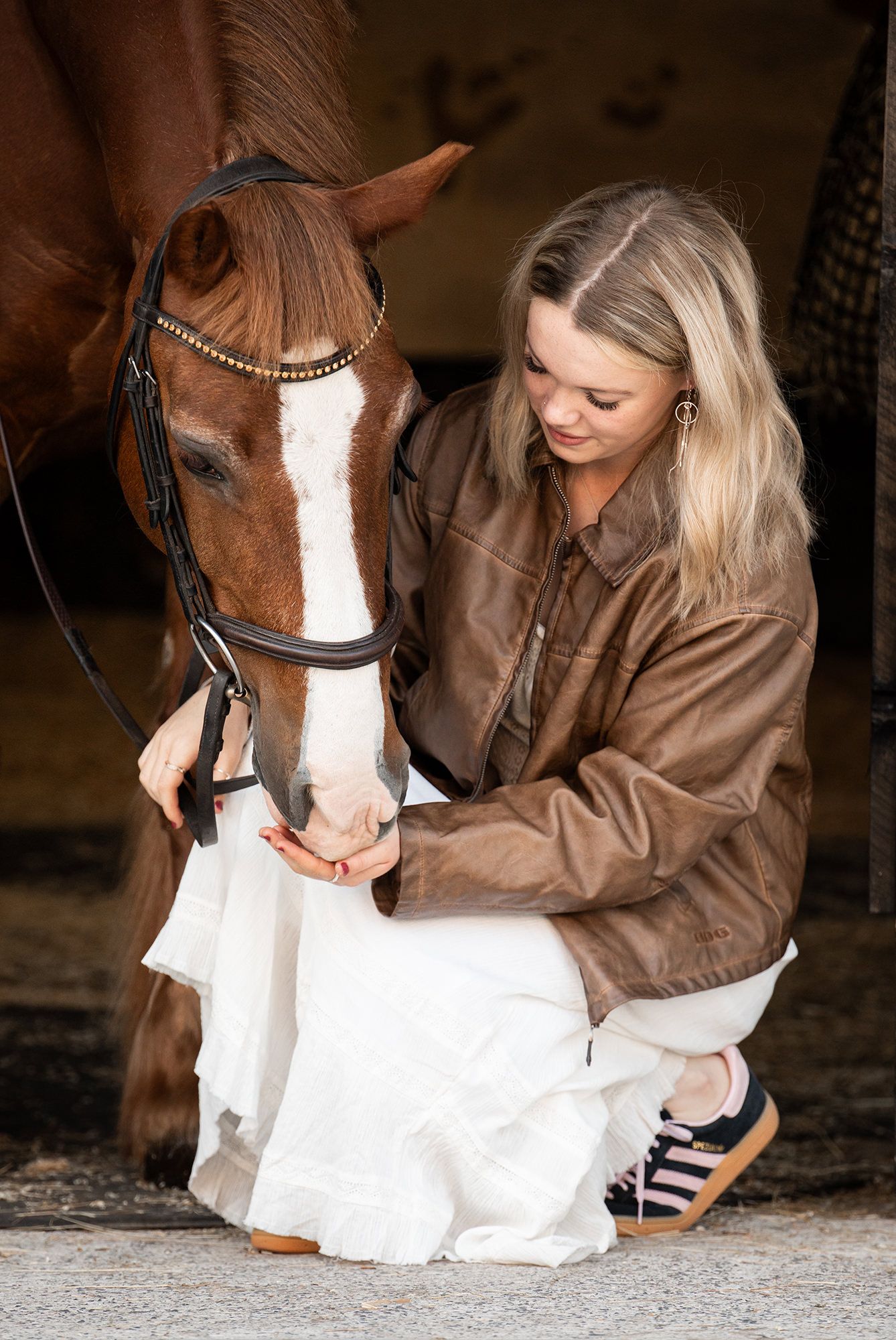 Sound girl in brown leather jacket tending to her horse with white facial markings in the stable at an equestrian property.