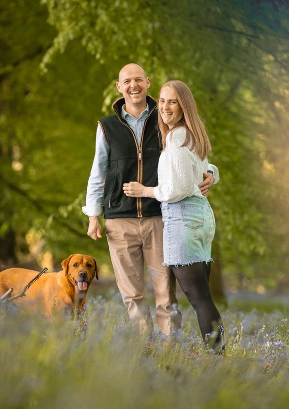A couple and their brown dog stand together in a field of bluebells during a sunny spring day.
