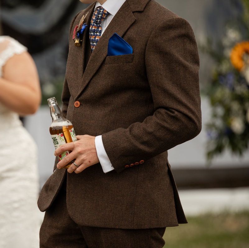 A brown tweed suit with blue pocket square and white shirt, holding a beer bottle at a wedding event.