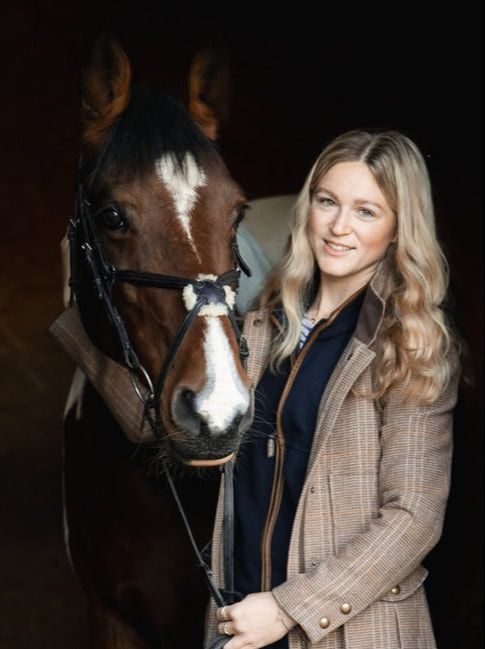 A bay horse with a white blaze stands next to a person wearing a brown plaid blazer in a stable setting.