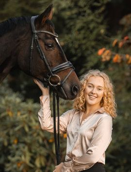 A brown horse with bridle stands next to someone in a beige shirt against a green foliage background.