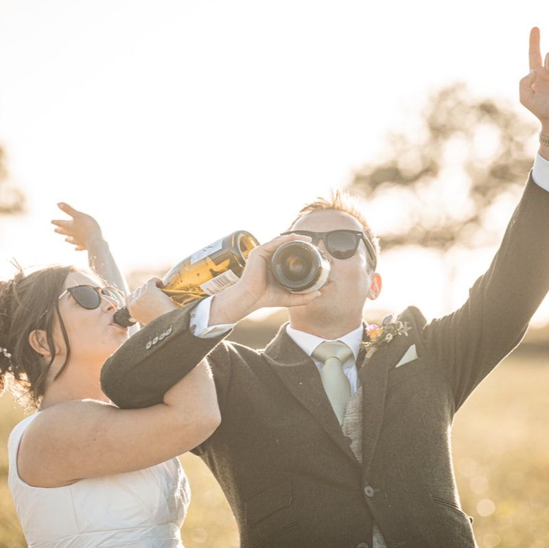 Wedding party celebrating with drinks and raised arms against golden sunset in outdoor celebration.