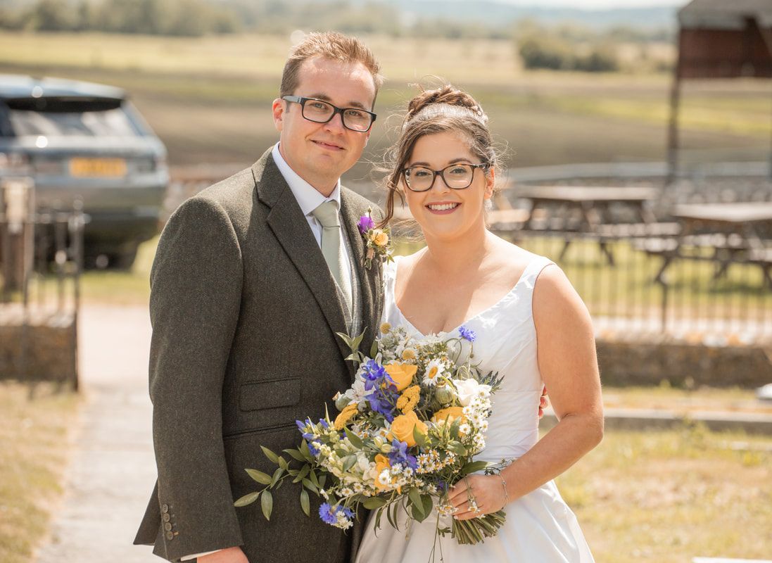 A happy couple in wedding attire poses outdoors with a bouquet of yellow and purple flowers on a sunny day.