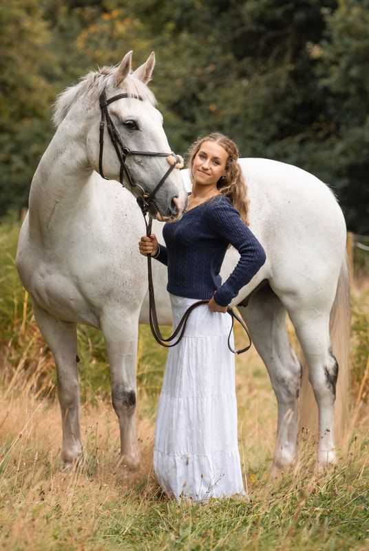 A beautiful white horse stands in a grassy field while being lovingly embraced in a tender outdoor photoshoot.