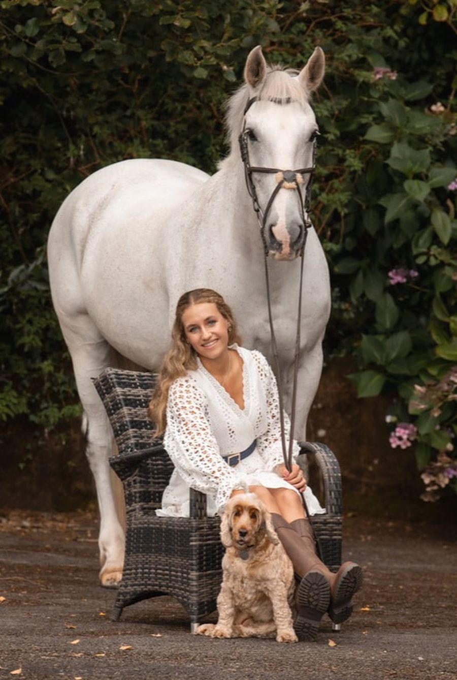 A white horse stands near a patio chair while a dog and someone in white sit together in a garden setting.