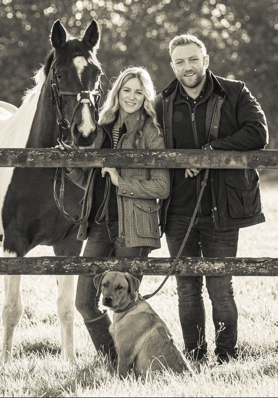 Black and white panoramic photo of a family with their horse standing by a wooden fence in a pastoral setting.