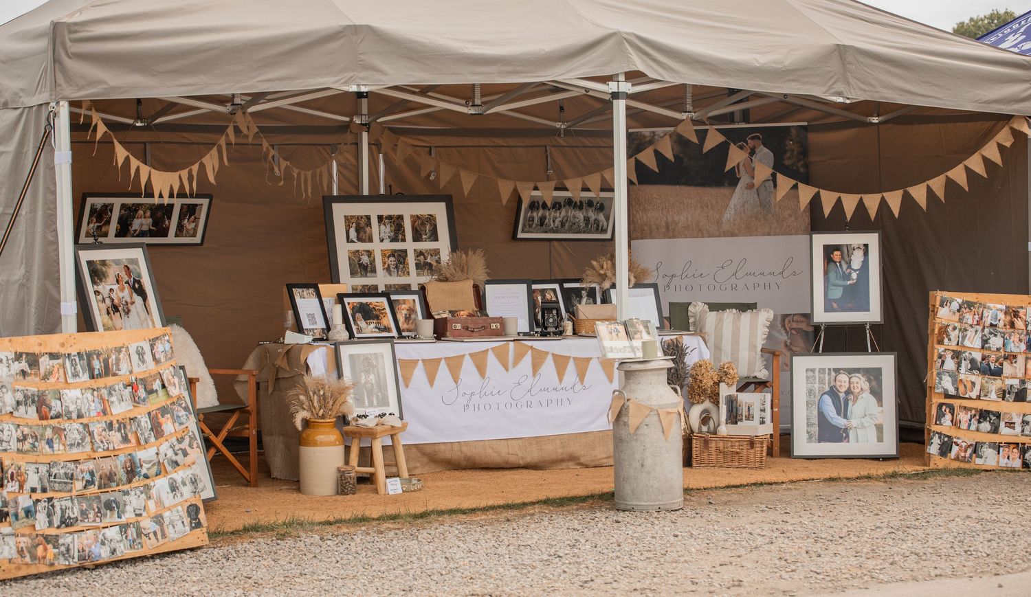 Rustic wedding display booth with framed photos, bunting, and decorative jars under a beige tent.