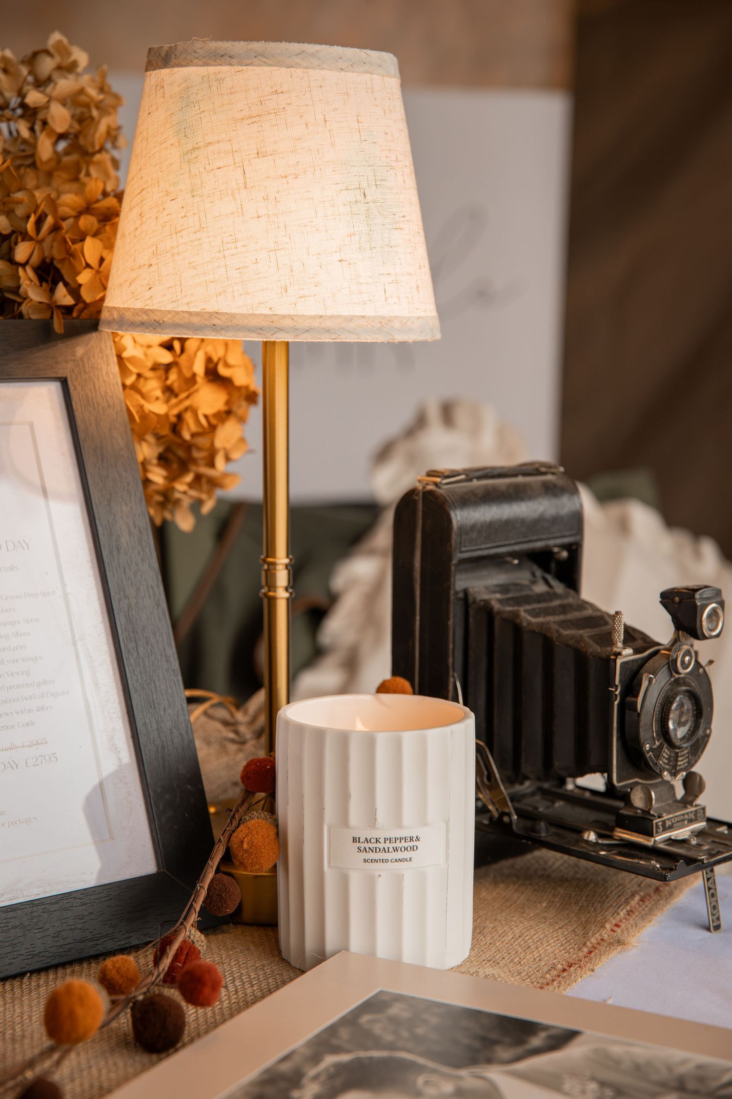 Vintage folding camera beside a white candle and brass table lamp with dried hydrangeas on a burlap surface.