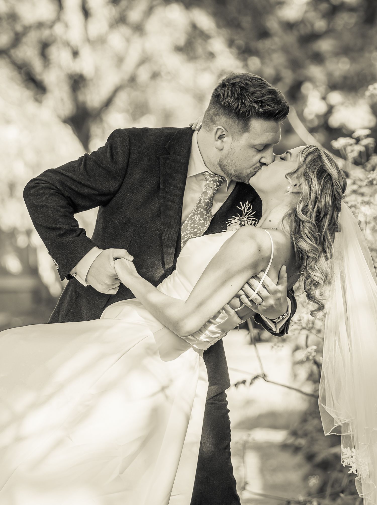Black and white photo of a groom dipping and kissing his bride in a romantic outdoor wedding portrait.