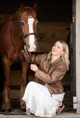 Person in brown jacket and white pants sitting on stable floor while grooming a horse's hooves.