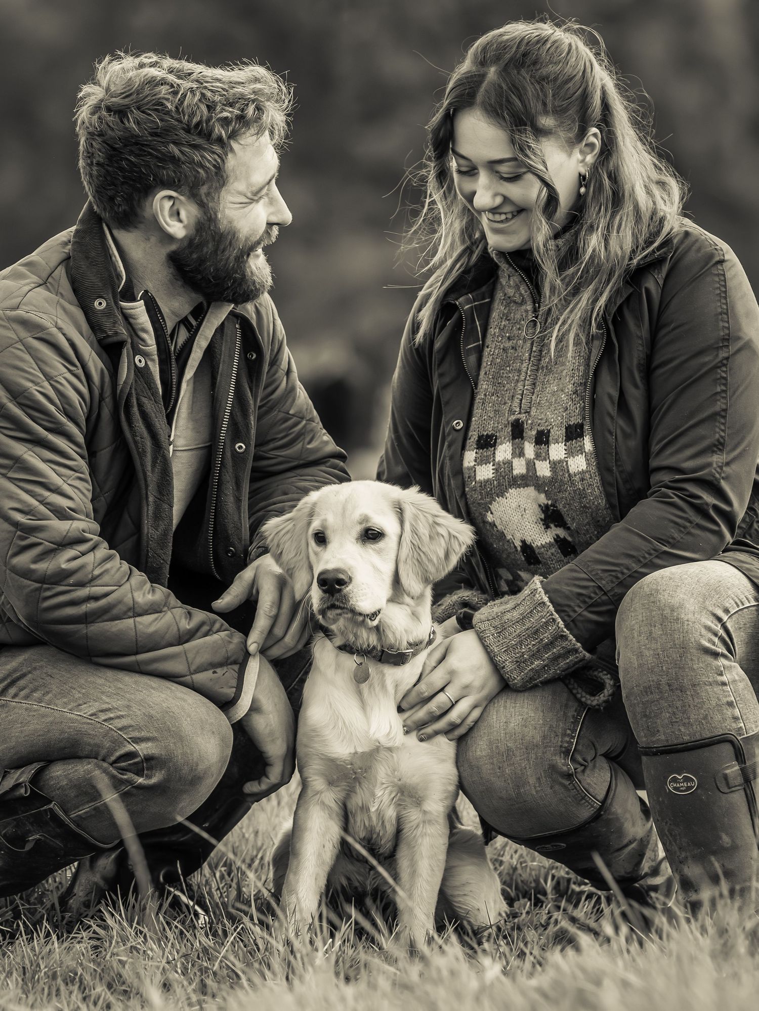 A couple sits on grass with their yellow Labrador retriever in a series of black and white engagement photos.