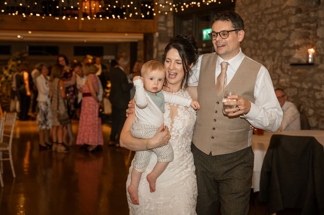 Wedding guests dance and celebrate on a rustic venue dance floor illuminated by warm string lights above.