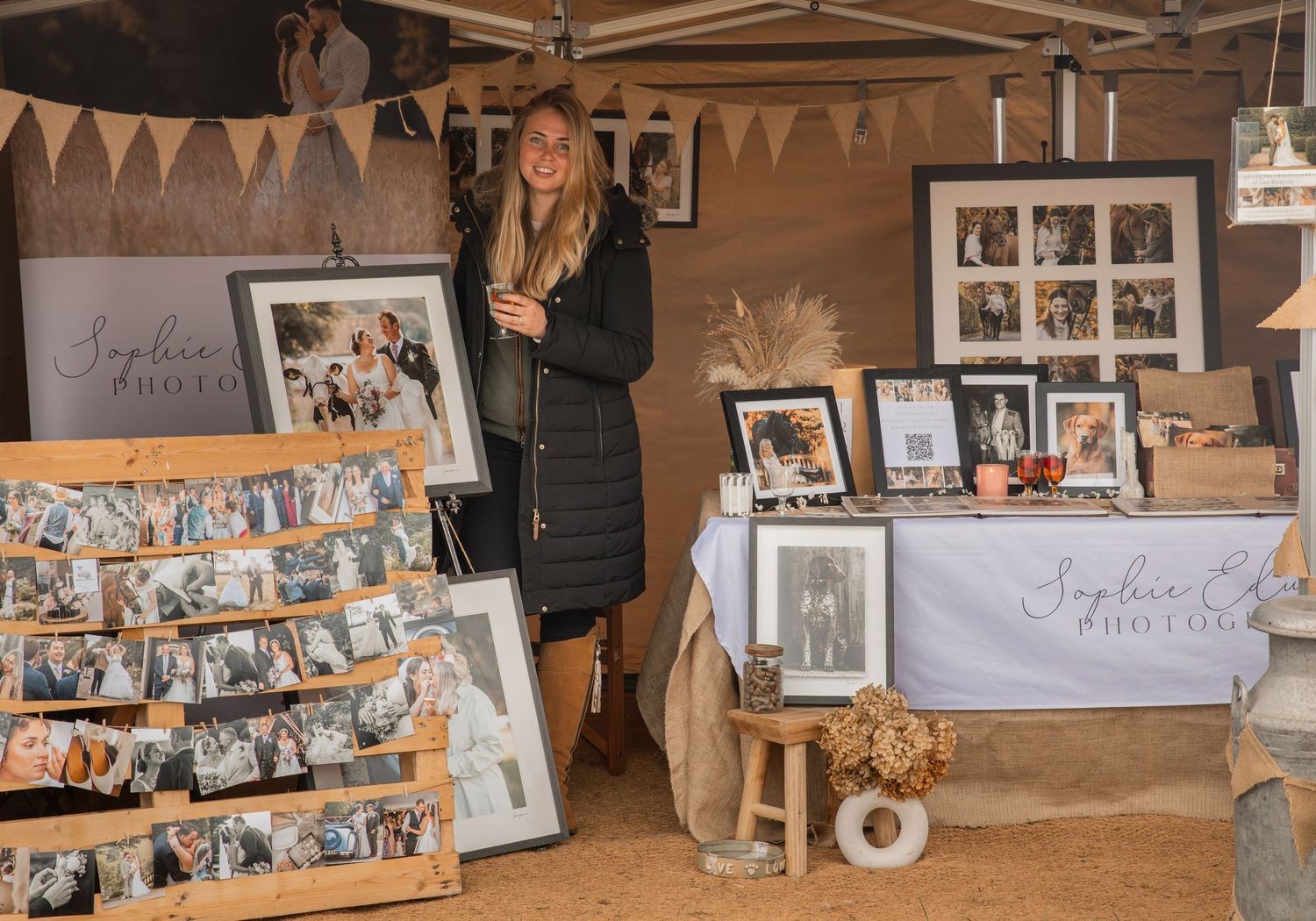 Woman stands at a photography display booth with framed prints and photo collections at an outdoor market event.