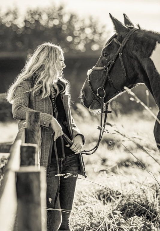 Black and white photo of someone in a winter coat standing beside a dark horse in a sunlit field.