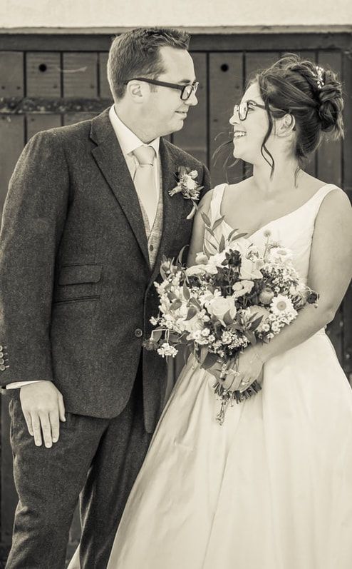 Black and white wedding photo showing couple in formal attire with glasses sharing a tender moment and bridal bouquet.