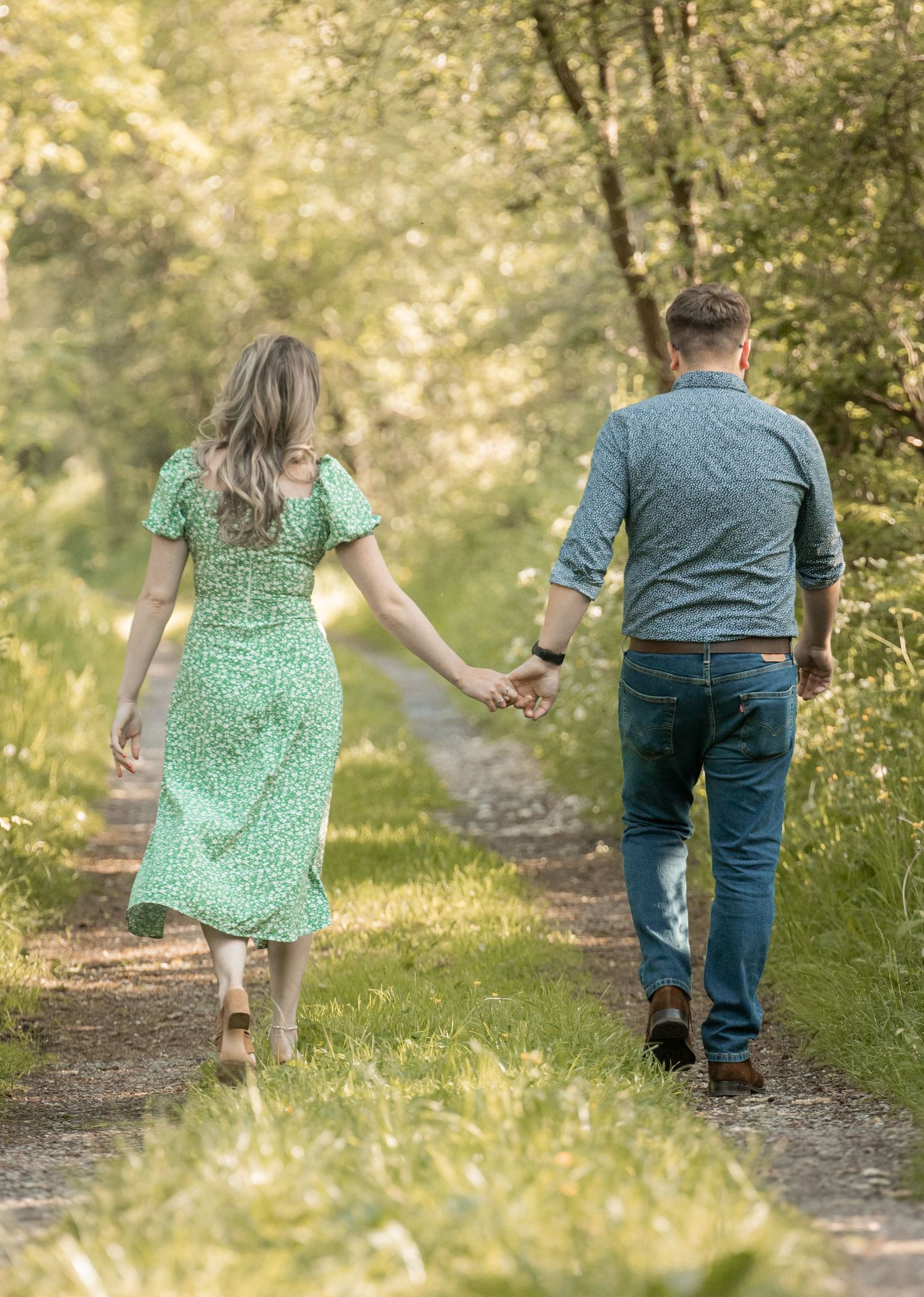 A couple walks hand in hand down a sunlit forest path wearing green and blue clothing on a warm summer day.