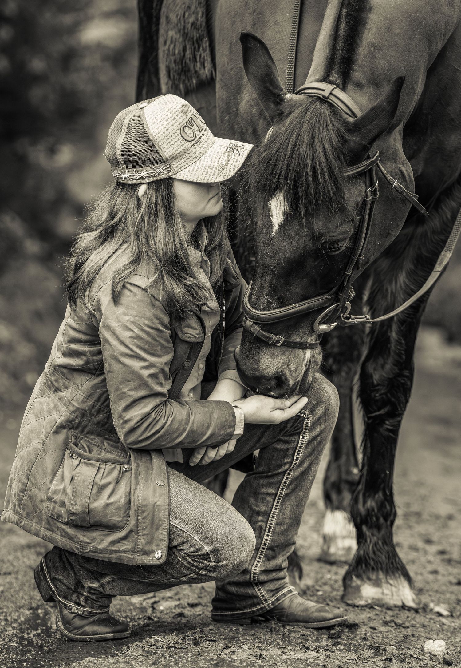 A black and white photo shows a person in a baseball cap and jacket kneeling beside a dark horse in rustic setting.