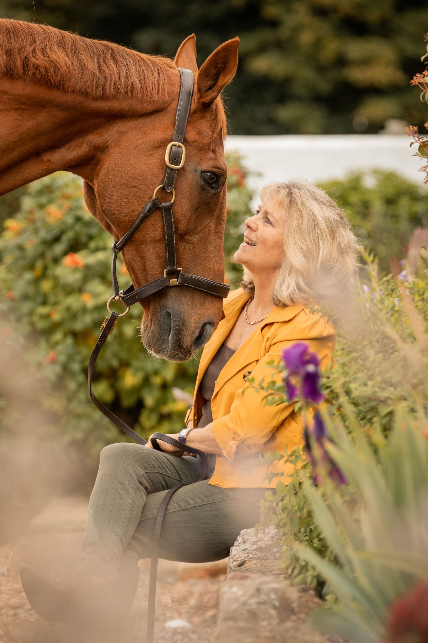 A person in a yellow jacket shares a tender moment with a chestnut brown horse in a garden setting.