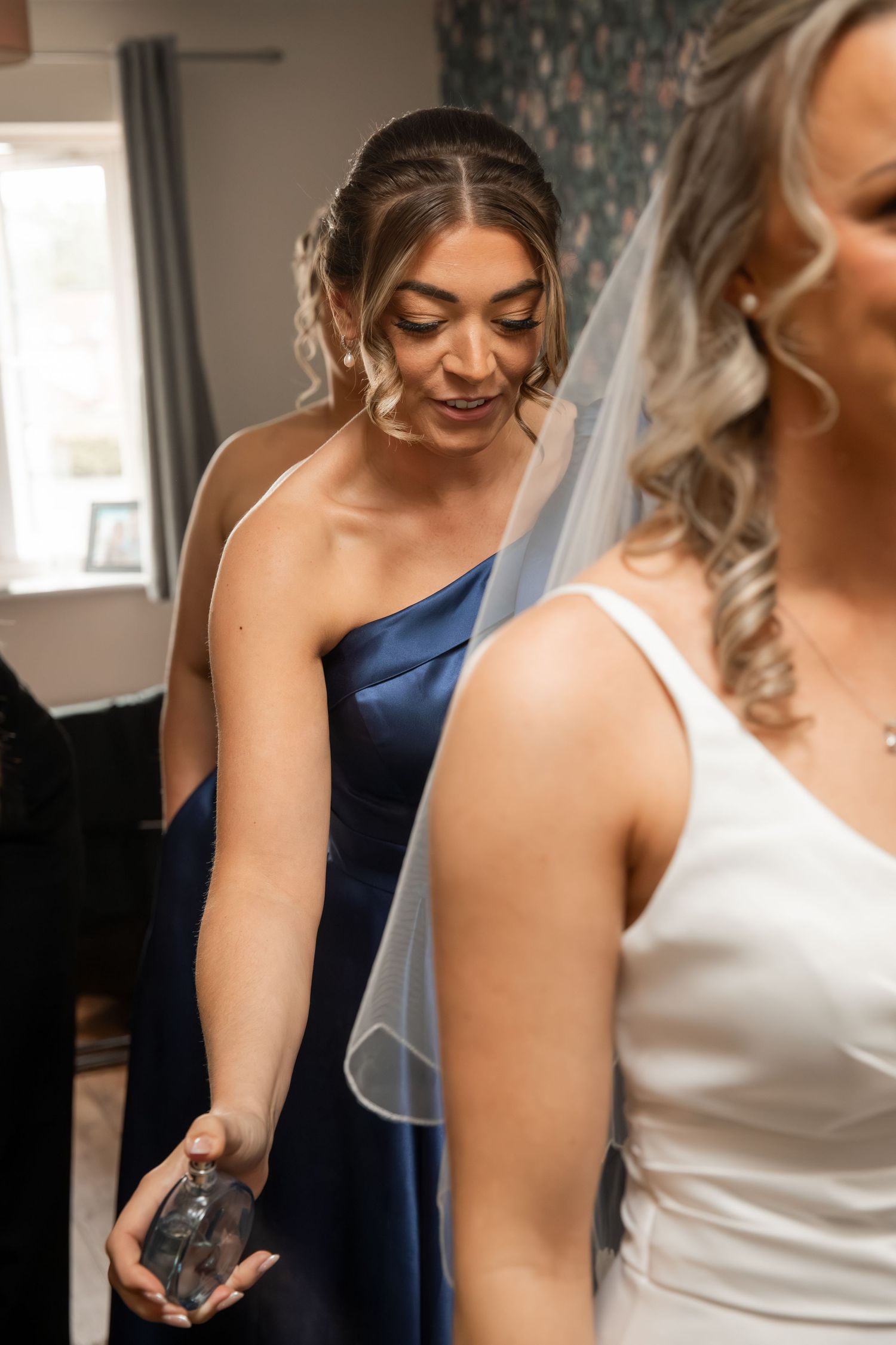 Bridesmaid in blue satin dress holding perfume bottle assists bride in white dress with veil during wedding preparations.