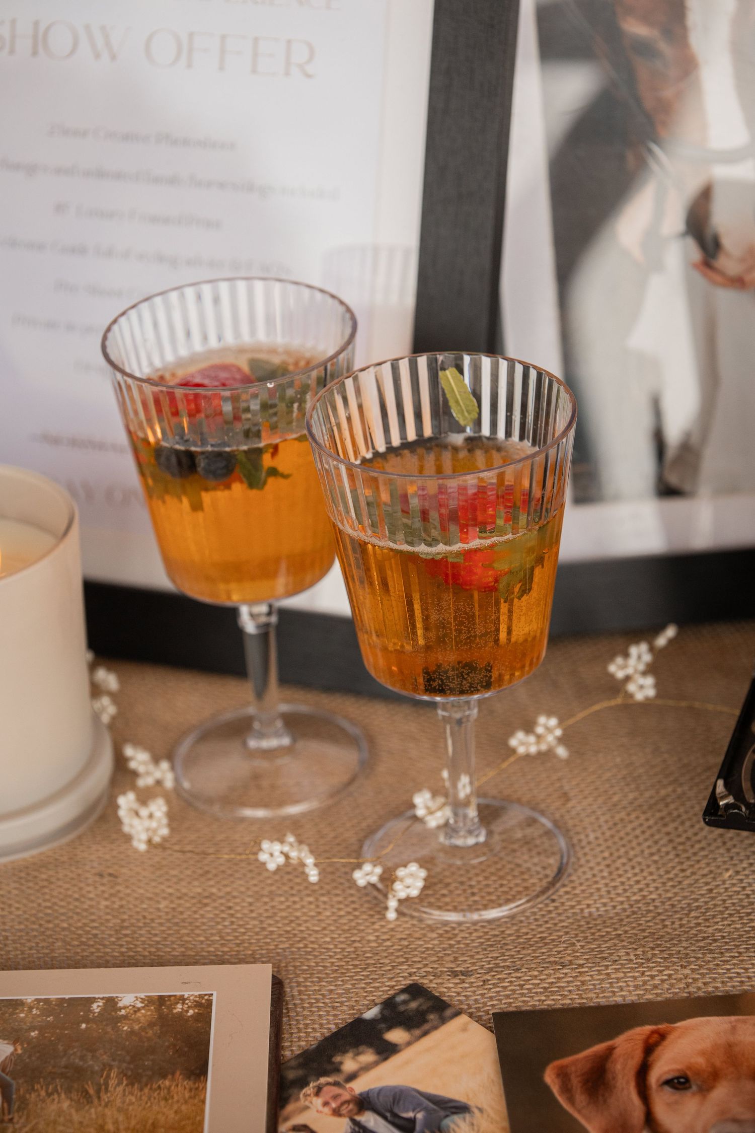 Two elegant ribbed glass goblets filled with amber cocktails garnished with fresh berries and mint leaves on a burlap surface.