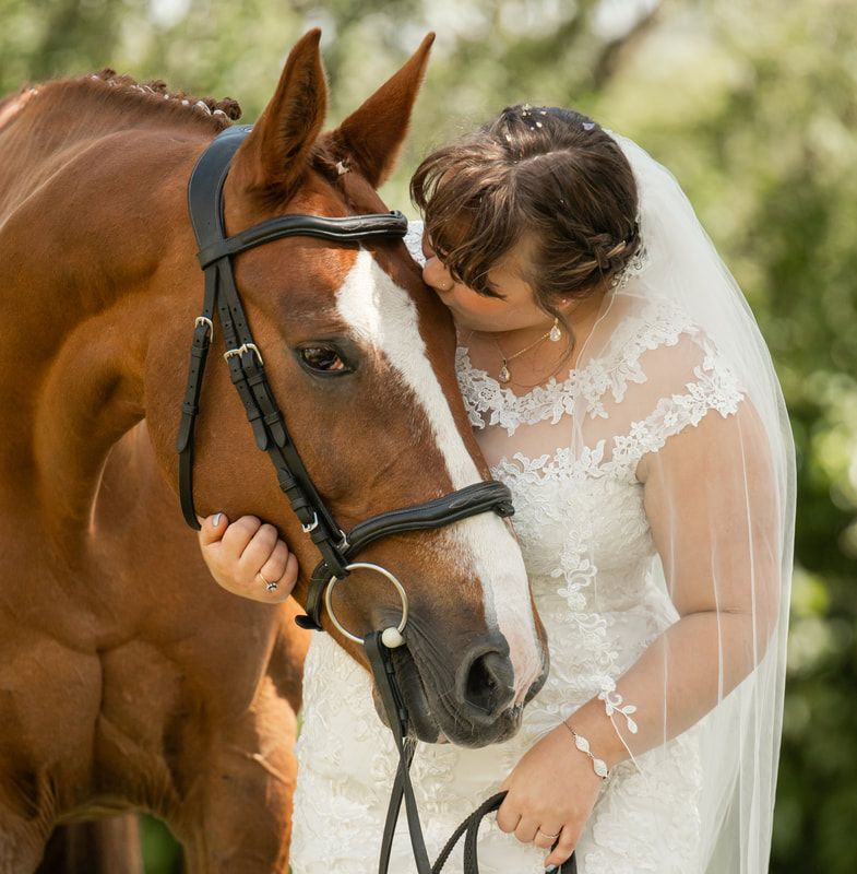 A bride in a white lace dress shares a tender moment with a brown horse wearing a black bridle in an outdoor setting.