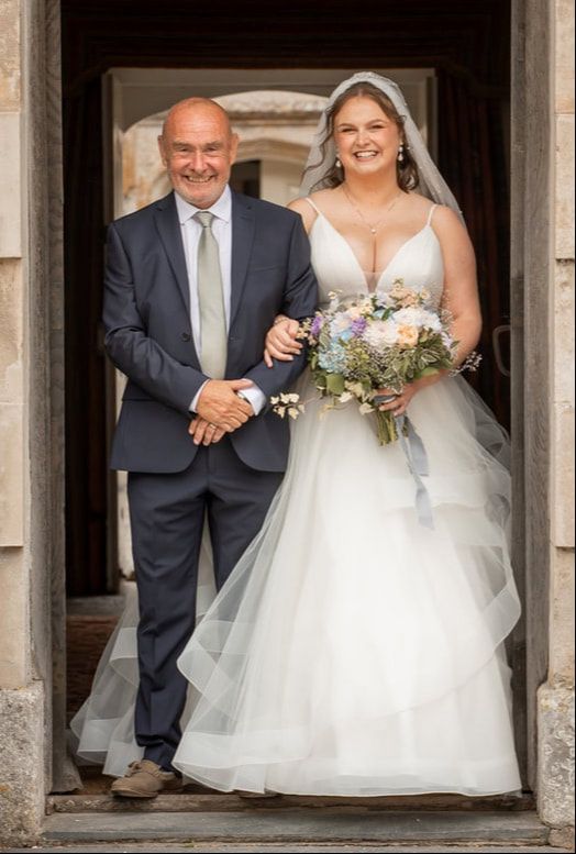 A bride in a white wedding dress stands with a companion in a navy suit in a stone doorway entrance.