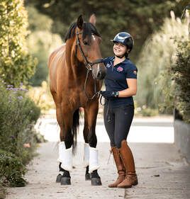 A rider in navy attire stands beside a chestnut horse on a sunlit outdoor path during an equestrian photoshoot.