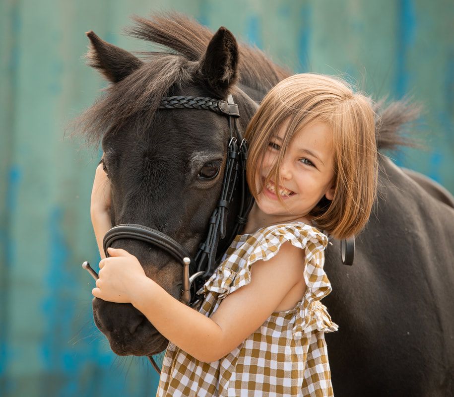 A young girl in a gingham dress hugs a black horse against a turquoise background.