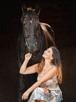 A dark horse and a woman in a floral dress share a tender moment against a dramatic black background.