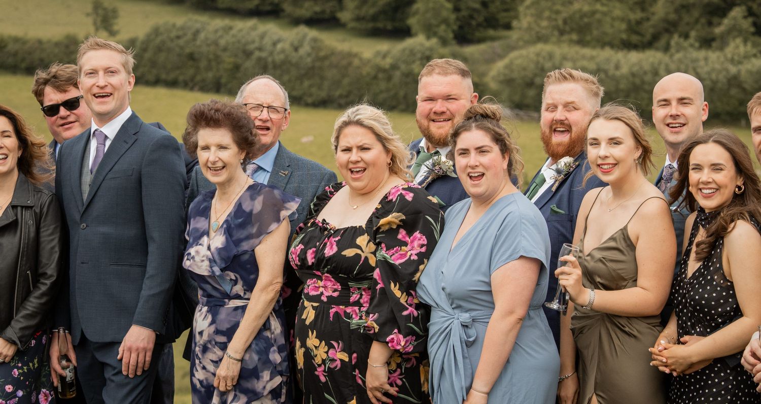 A group of wedding guests share joyful laughter while standing together outdoors.