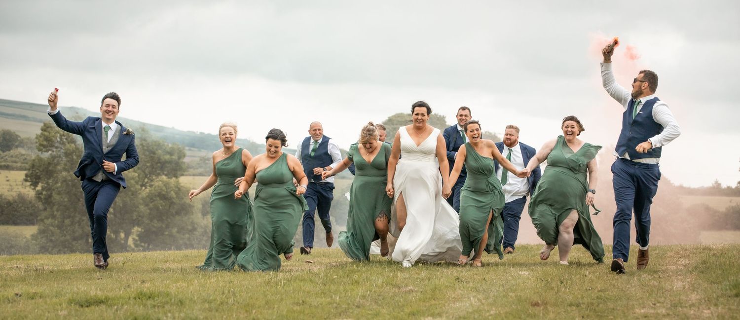 Wedding party in green bridesmaid dresses and navy suits jumps joyfully on a grassy hillside under cloudy skies.