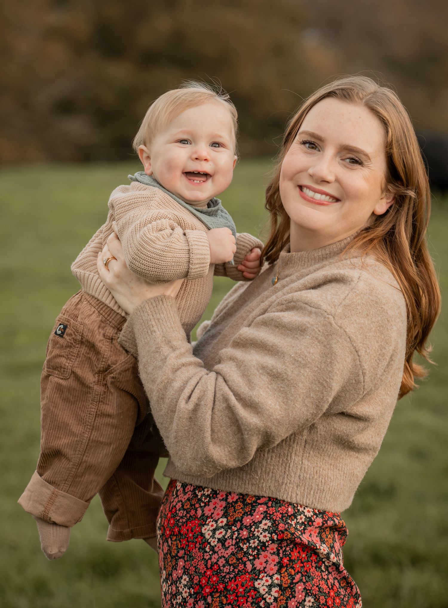 A mother and baby share joyful moments together outdoors in warm earth-toned sweaters and floral dress.