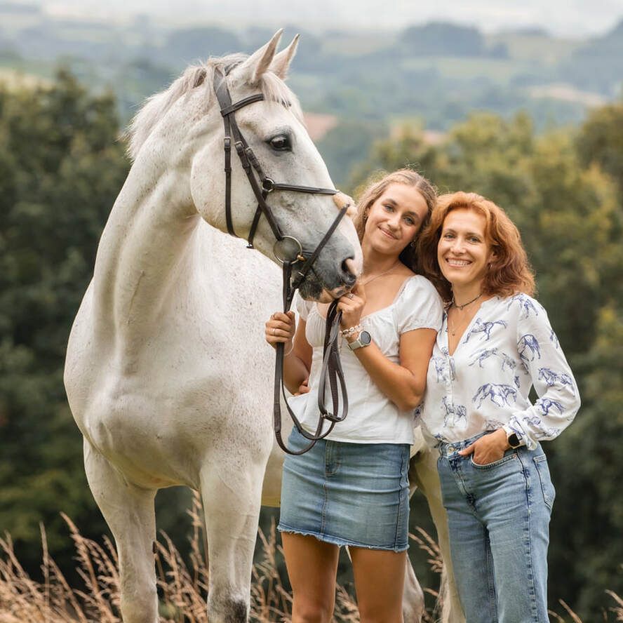 Beautiful white horse stands with two people in casual wear against a scenic natural backdrop on a sunny day.