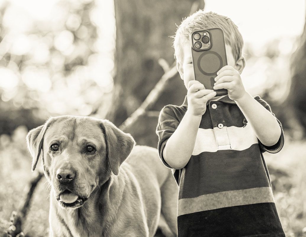 Black and white photo of a child taking a picture with a smartphone next to a curious yellow Labrador retriever.