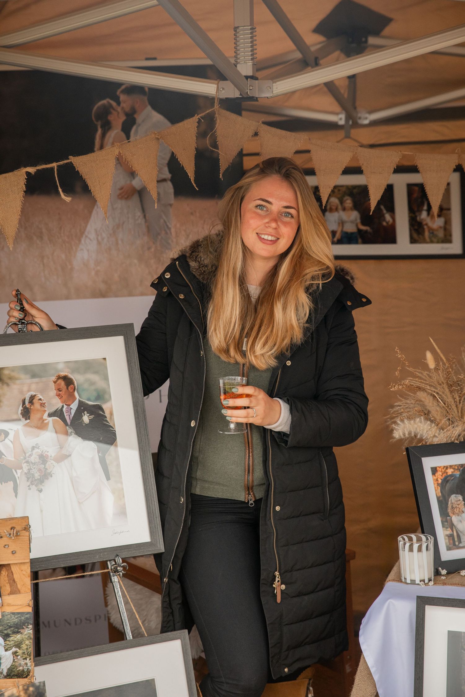 Woman in black puffer jacket holding a drink at a wedding photography exhibition with framed prints on display.