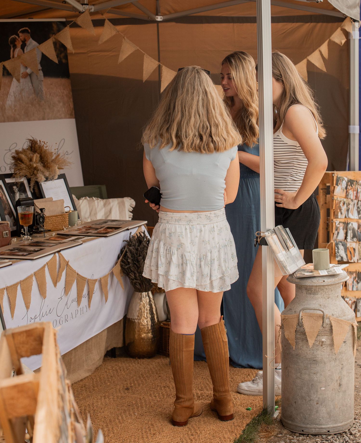 Two women browse a rustic wedding vendor booth with burlap decor, vintage milk can, and displayed photos at an outdoor market.